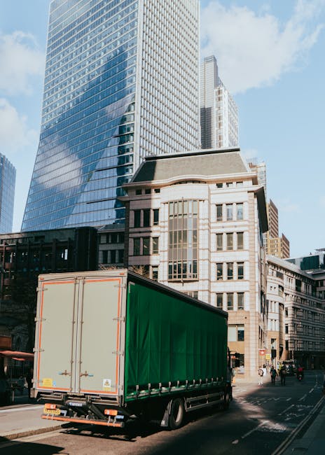 A large moving truck with a green cargo area is parked on a city street in front of tall modern office buildings with glass facades and a historic multi-storey building with stone details. The truck is positioned near the curb, with its rear doors closed, and is prepared for a house removal or furniture transport. The scene shows a clear day with blue skies and some sunlight reflecting off the buildings. Pedestrians and cyclists are visible along the sidewalk, and the traffic appears light. The image captures the process of home relocation in an urban environment, with the truck ready for loading or unloading furniture, boxes, and packing materials as part of a professional removals service by Man With a Van Haringey.