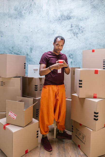 A man in a maroon t-shirt and bright orange trousers stands in front of a light grey textured wall, surrounded by numerous sealed cardboard boxes of various sizes with red and black tape, some open revealing packing materials. He is holding a smartphone with a red case in both hands, looking directly at the camera. The boxes are stacked on the floor, some partially blocking the pathway, indicating a packing or home relocation process. The setting appears to be indoors, likely a storage area or part of a house during packing for a move. The scene reflects the packing and moving preparations associated with professional removals services, such as those offered by Man With a Van Haringey.