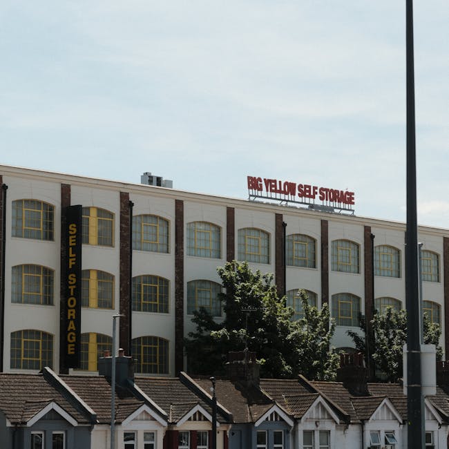 A large moving truck with a green cargo area is parked on a city street in front of tall modern office buildings with glass facades and a historic multi-storey building with stone details. The truck is positioned near the curb, with its rear doors closed, and is prepared for a house removal or furniture transport. The scene shows a clear day with blue skies and some sunlight reflecting off the buildings. Pedestrians and cyclists are visible along the sidewalk, and the traffic appears light. The image captures the process of home relocation in an urban environment, with the truck ready for loading or unloading furniture, boxes, and packing materials as part of a professional removals service by Man With a Van Haringey.