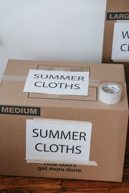 A close-up view of a cardboard moving box placed on a wooden floor, used for home relocation or packing during house removals. The box features printed labels indicating its contents, specifically 'SUMMER CLOTHS' with one label attached with clear tape on the front and another label on top of the box, both in bold black text. Additionally, a white roll of packing tape with a partially visible brand name is positioned on the top of the box. In the background, part of another cardboard box with similar labels and size indicators is visible, suggesting the packing process is underway. The scene is well-lit, showing typical moving equipment used by companies like Man With a Van Haringey during furniture transport and packing and moving services, highlighting the importance of proper organisation in the loading process for a smooth house removal.