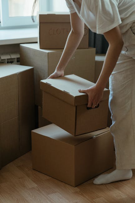 A close-up view of a cardboard moving box placed on a wooden floor, used for home relocation or packing during house removals. The box features printed labels indicating its contents, specifically 'SUMMER CLOTHS' with one label attached with clear tape on the front and another label on top of the box, both in bold black text. Additionally, a white roll of packing tape with a partially visible brand name is positioned on the top of the box. In the background, part of another cardboard box with similar labels and size indicators is visible, suggesting the packing process is underway. The scene is well-lit, showing typical moving equipment used by companies like Man With a Van Haringey during furniture transport and packing and moving services, highlighting the importance of proper organisation in the loading process for a smooth house removal.
