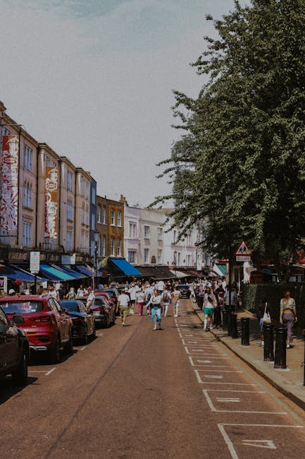 A busy street scene during market day showing parked cars along the curb and a line of pedestrians walking on the pavement, some carrying bags or pushing strollers. The street is lined with colorful, multi-story buildings with shopfronts and awnings. A large tree with dense foliage extends over part of the sidewalk, providing shade. In the foreground, the surface of the street is visible with marked parking bays and a tactile paving strip. The environment appears vibrant and lively, with the street likely experiencing high footfall. For home relocation or furniture transport purposes, this scene highlights the importance of planning routes to avoid crowded area congestion during market days, as emphasized by [COMPANY_NAME] in their guidance on routes to avoid on market days at Crouch End, Haringey.