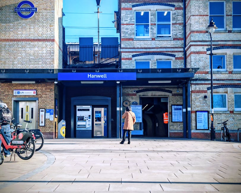 A woman dressed in a beige coat and dark trousers stands on the paved entrance area of Hanwell underground station, part of the Elizabeth Line, in Haringey. She appears to be looking at the station’s ticket machines, which are situated beneath a dark canopy with the station name displayed on a blue sign. Adjacent to the ticket machines, there is a row of bicycles and e-scooters parked on the left side, including bikes branded with Uber and VANS logos. The station building has a multi-storied brick exterior with several windows, some featuring small balconies with black railings. To the right, a narrow passageway leads into the underground station, with an orange safety vest visible on a person inside. The scene is lit by a traditional street lamp attached to the building, with bright daylight casting shadows on the pavement, capturing an everyday moment of home relocation preparations or transit organization, relevant to moving and transport logistics supported by Man With a Van Haringey.