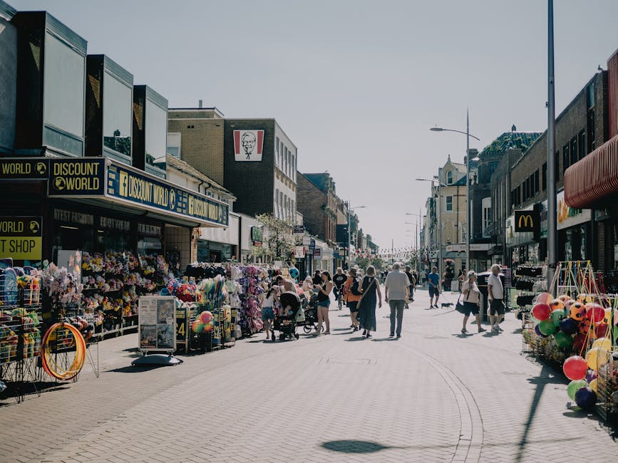 A busy street scene during market day showing parked cars along the curb and a line of pedestrians walking on the pavement, some carrying bags or pushing strollers. The street is lined with colorful, multi-story buildings with shopfronts and awnings. A large tree with dense foliage extends over part of the sidewalk, providing shade. In the foreground, the surface of the street is visible with marked parking bays and a tactile paving strip. The environment appears vibrant and lively, with the street likely experiencing high footfall. For home relocation or furniture transport purposes, this scene highlights the importance of planning routes to avoid crowded area congestion during market days, as emphasized by [COMPANY_NAME] in their guidance on routes to avoid on market days at Crouch End, Haringey.