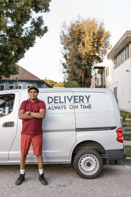 A young male delivery driver stands confidently with arms crossed in front of a white delivery van parked outside a residential property during daylight. The van displays the words 'DELIVERY ALWAYS ON TIME' on its side, indicating a professional transportation service. The driver is dressed in a red polo shirt, orange shorts, and black sports shoes, with a cap on his head. The background features trees with autumn foliage, a modern white building with large windows, and a partly cloudy sky. The scene suggests a home relocation or furniture transport context, with the van parked near the property's entrance, ready for packing and moving tasks, and the driver prepared for efficient delivery or removal services, as offered by Man With a Van Haringey.