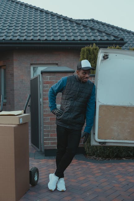 A young man with a beard, wearing a black cap and a white collared shirt, standing inside a loading van during a home relocation process. The van's open rear doors reveal numerous cardboard boxes, some wrapped in plastic film, stacked neatly inside. The man is positioned in front of the boxes with his arms crossed, facing the camera with a serious expression. The environment is well-lit, and the interior of the van is visible, showing various packed items ready for transportation. The scene depicts the process of furniture transport and packing during a professional removal service, with the van situated on a pavement area outside a residential property, supporting the context of removal and moving logistics associated with Man With a Van Haringey.