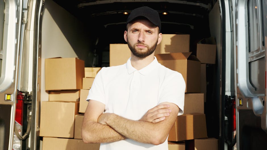 A man in a white polo shirt and black cap standing inside a cargo van used for house removals, with his arms crossed and a serious expression. The van's interior is visible behind him, filled with numerous cardboard boxes of various sizes, some sealed with packing tape and others open, revealing packing materials like bubble wrap and plastic wrapping. The van doors are open, showing the pavement outside, indicating the loading or unloading process during a furniture transport or home relocation. Man With a Van Haringey is providing furniture transport services, and the image highlights the careful handling of packed items for house removals or moving logistics, emphasizing the loading process and use of moving equipment such as trolleys or blankets.
