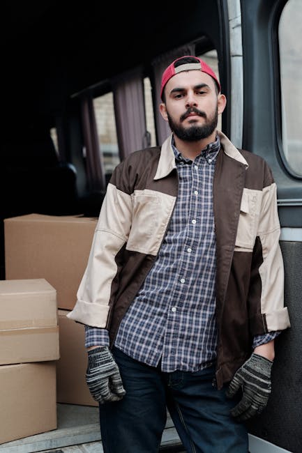 A man with a beard and dark hair, wearing a red backward cap, a brown and beige jacket over a blue and white checked shirt, and grey work gloves, stands inside the back of a van during a home relocation. The van's interior shows cardboard boxes of various sizes packed with packing materials, ready for transport. The man is positioned near the doorway, with the van's interior partially visible behind him, indicating the loading process involved in furniture transport and packing for a move, consistent with the services of Man With a Van Haringey. Natural lighting highlights the scene, emphasizing the professional organization involved in house removals and moving logistics.
