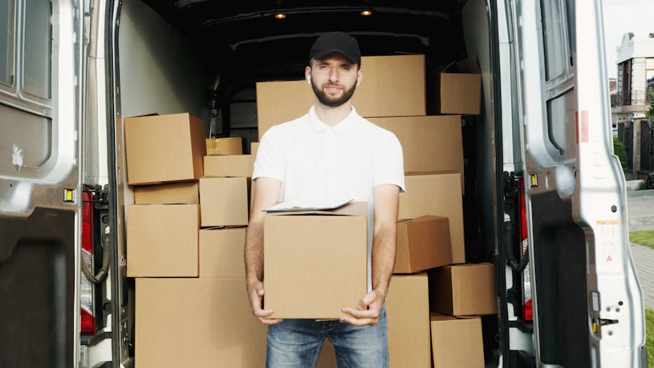 A man in a white polo shirt and black cap standing inside a cargo van used for house removals, with his arms crossed and a serious expression. The van's interior is visible behind him, filled with numerous cardboard boxes of various sizes, some sealed with packing tape and others open, revealing packing materials like bubble wrap and plastic wrapping. The van doors are open, showing the pavement outside, indicating the loading or unloading process during a furniture transport or home relocation. Man With a Van Haringey is providing furniture transport services, and the image highlights the careful handling of packed items for house removals or moving logistics, emphasizing the loading process and use of moving equipment such as trolleys or blankets.