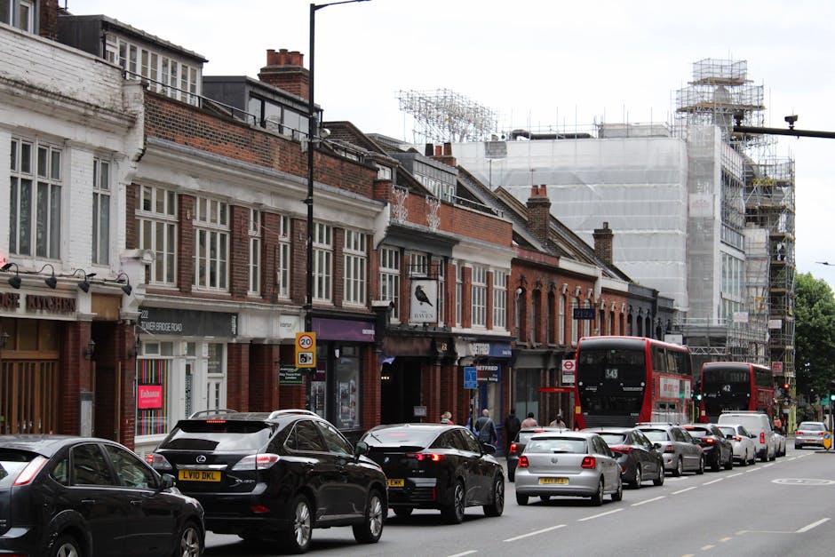 A street scene in Haringey showing a row of brick and white-painted commercial buildings with large windows, some with signage, and a scaffolding-covered building under construction in the background. On the pavement, several pedestrians walk past parked and moving vehicles, including a white van partially visible on the left, and multiple cars parked along the curb. Two red double-decker buses are in motion, one near the centre and another further down the street. The road is filled with cars and SUVs, some waiting as another bus approaches. Street signs indicating speed limits and parking restrictions are visible. The scene appears during the day with overcast weather, and the environment suggests active home and business relocation activities, with the presence of vehicles used for furniture transport and moving equipment, supporting the context of house removals and street loading permits facilitated by [COMPANY_NAME].
