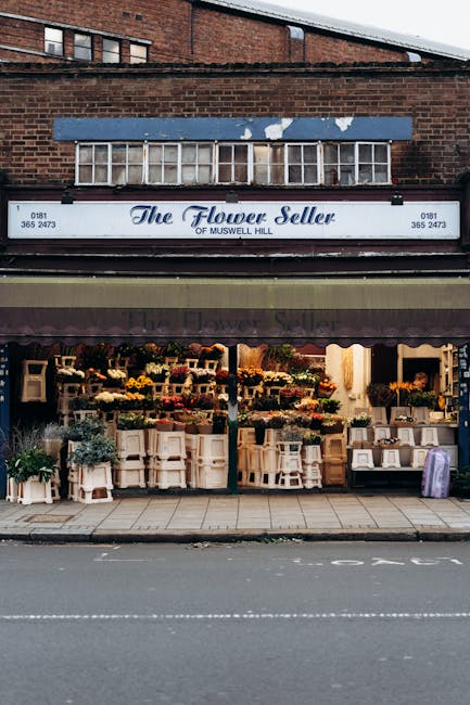The image depicts the storefront of a flower shop called 'The Flower Seller of Muswell Hill,' situated on a city street. The shop has a brown awning with the store's name displayed in white lettering, partially shading the display area. In front of the shop, there are numerous flower arrangements and potted plants arranged on white shelving units and tables, showcasing a variety of colourful flowers and greenery. The shop's large glass windows reveal more floral displays inside, and a faint warm interior light is visible. Above the shop, a sign indicates street loading rules during relocations or removals in Haringey, with a brick building and small window above the sign. The pavement in front of the shop is clean, and the street appears calm and quiet, suitable for loading or unloading when parking restrictions permit. Occasionally, [COMPANY_NAME] and similar services may use such locations for furniture transport and home relocation activities, ensuring minimal disruption to local businesses and residents.