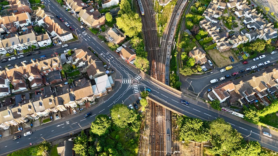 An aerial view of a residential neighbourhood showing a busy intersection with multiple roads, parked cars, and houses with pitched roofs and gardens. The image captures a railway bridge crossing over the roads, with visible train tracks and overhead electrical lines. Surrounding the streets are green trees and well-maintained gardens, indicating a suburban area. The scene is taken during daylight with clear weather, highlighting the urban layout and infrastructure that may present challenges during a house relocation or furniture transport process. This setting underscores common access issues faced during home removals in the N15 area, where careful planning for parking, street access, and bridge clearance is essential, as managed by companies like Man With a Van Haringey.