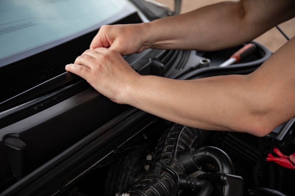 Close-up image showing a person's hands inspecting and adjusting the engine bay of a vehicle, with the hood open. The engine components, including black hoses and metal parts, are visible beneath the hood. The person’s arms are positioned inside the engine compartment, working on the area near the engine cover. The environment appears to be a well-lit garage or driveway setting. This scene illustrates the preparatory steps involved in vehicle inspection during a home relocation or furniture transport process, possibly handled by Man With a Van Haringey. The focusing on engine checks aligns with the logistical considerations of moving your belongings safely and efficiently.