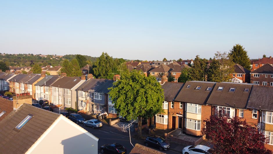 An aerial view of a residential neighbourhood showing a busy intersection with multiple roads, parked cars, and houses with pitched roofs and gardens. The image captures a railway bridge crossing over the roads, with visible train tracks and overhead electrical lines. Surrounding the streets are green trees and well-maintained gardens, indicating a suburban area. The scene is taken during daylight with clear weather, highlighting the urban layout and infrastructure that may present challenges during a house relocation or furniture transport process. This setting underscores common access issues faced during home removals in the N15 area, where careful planning for parking, street access, and bridge clearance is essential, as managed by companies like Man With a Van Haringey.