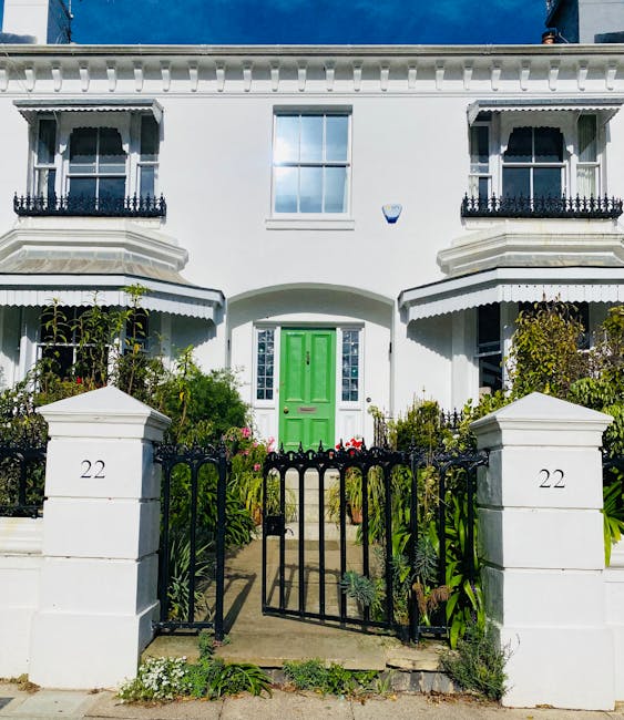 The image shows the front view of a white two-storey residential building located at number 22, with a bright green front door centered above a small concrete pathway. The house features decorative black wrought iron balcony railings on two upper-story windows, and the structure has crisp architectural details such as corner pilasters and a decorative cornice. In front of the property, a black metal gate with a simple design opens onto a paved pathway leading to the entrance, flanked by two white brick pillars also marked with the number 22. Surrounding the pathway, there are well-maintained green plants, shrubs, and small flowering bushes, indicating a garden area. The overall lighting suggests bright daylight conditions, with some shadows cast on the building façade. This scene could relate to house removals or home relocation processes managed by Man With a Van Haringey, especially in the context of packing and preparation for a move in the N22 area of Wood Green.