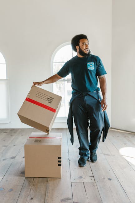 A man and woman indoors, preparing for a home relocation, both holding moving boxes; the man, wearing an orange pair of trousers and a maroon t-shirt, is smiling and holding a small cardboard box with a 'Fragile' label in his left hand, while the woman, in a white top and blue jeans, is seated on a green sofa and holds a larger cardboard box. They are positioned near a large window with natural light illuminating the room, which features potted plants, including a large leafy one in a white textured vase, and wooden flooring. Their relaxed expressions suggest a collaborative packing or unpacking process, as part of a furniture transport or packing and moving operation. The scene reflects a casual, organized approach to house removals, with visible cardboard boxes, interior decor, and natural environment, consistent with professional removals services such as those offered by Man With a Van Haringey specializing in local house and flat removals within the N22 area.